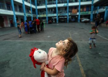 Aya, a 5-year-old girl clutching her doll to ease her fear, gazing at Gaza's sky filled with warplanes from inside an UNRWA school in the Gaza Strip.