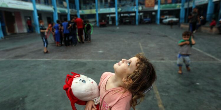 Aya, a 5-year-old girl clutching her doll to ease her fear, gazing at Gaza's sky filled with warplanes from inside an UNRWA school in the Gaza Strip.